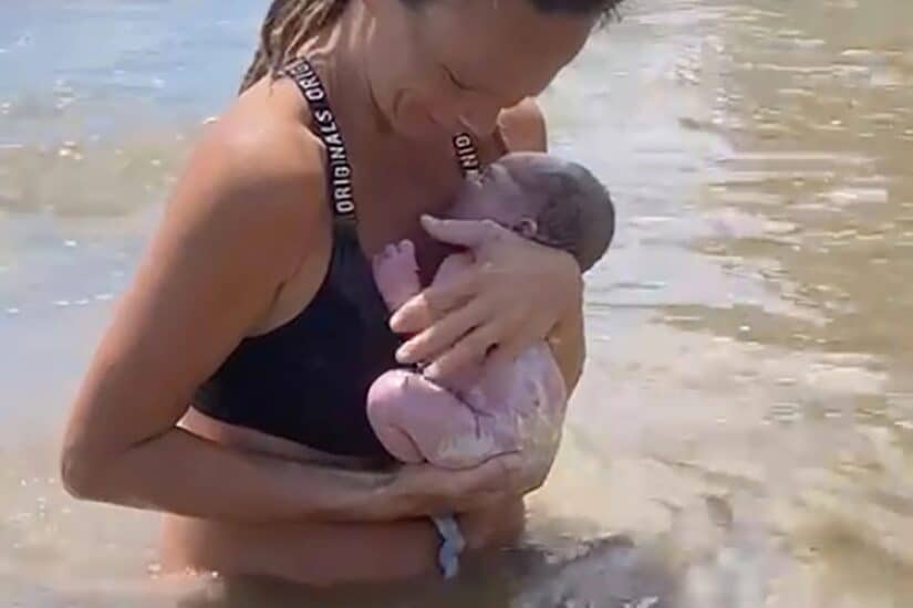 Mother holding newborn baby in shallow water at the beach.