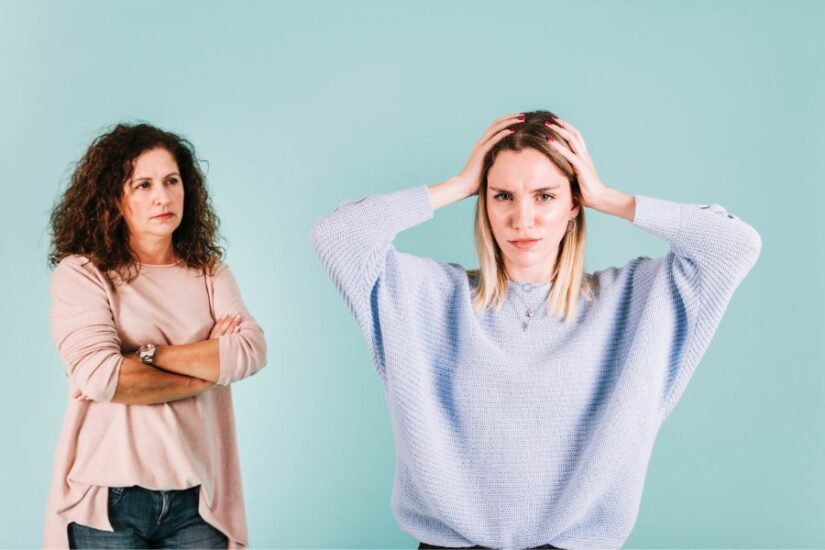 An upset woman holding her head in frustration with another woman standing nearby with crossed arms.