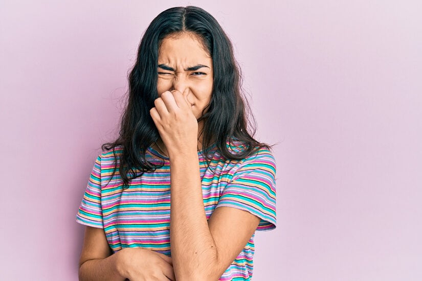 Young woman covering her nose, showing bad breath discomfort, on pink background.
