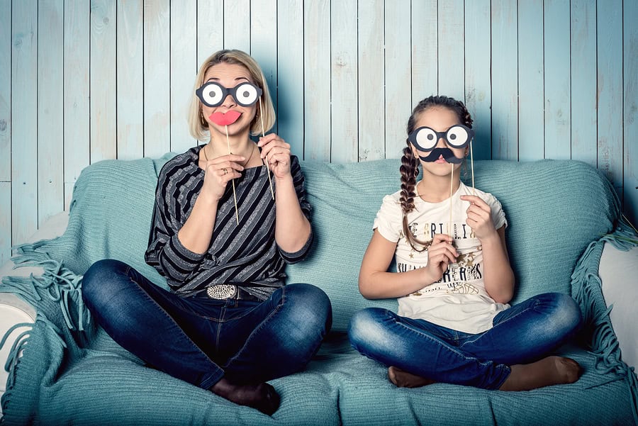 Playful mom and daughter wearing humorous masks, sitting on a comfy sofa at home.