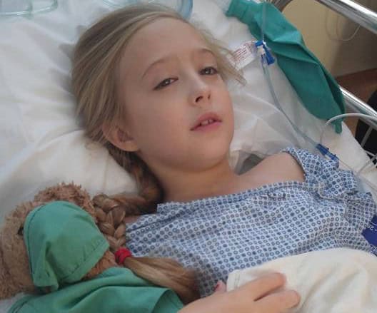 Young girl lying in hospital bed with medical tubes, battling breast cancer at age 8.