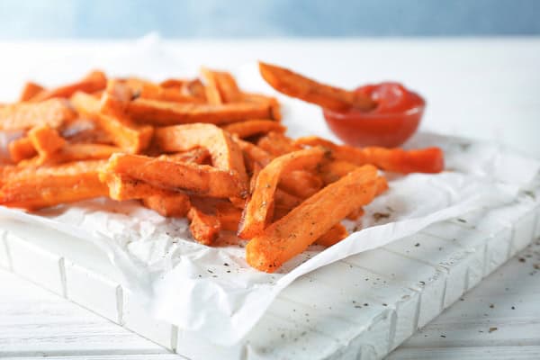 Crispy baked sweet potato fries with a side of ketchup on white parchment paper.