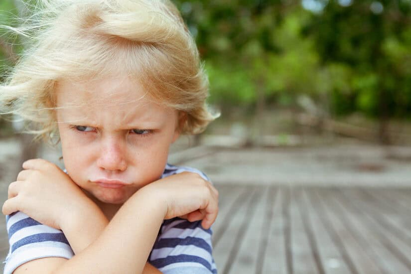 Child with a frustrated expression, arms crossed, outdoors on a wooden deck.