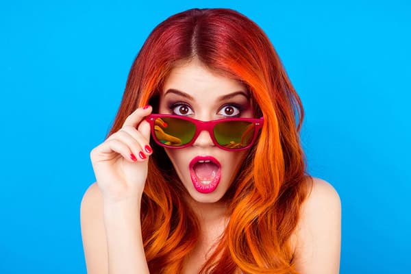 Surprised woman with red hair and sunglasses on blue background.