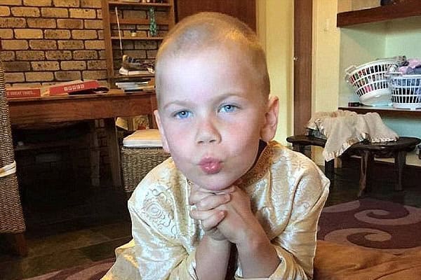 Young boy with cancer sitting with hands clasped, thoughtful expression.