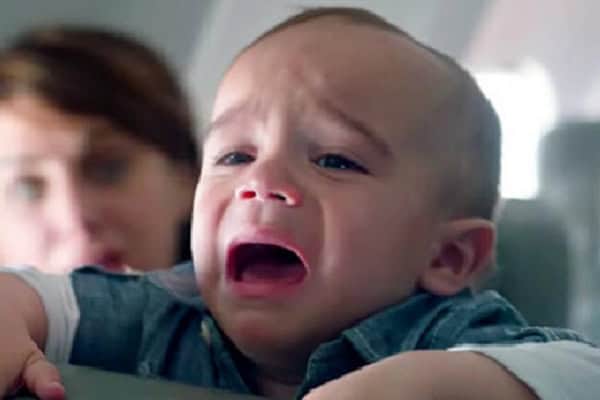Baby crying during flight, distressed child with mother in background.