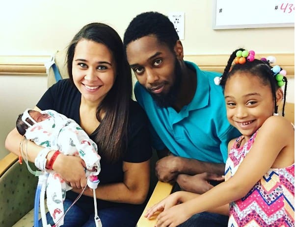Baby born in amniotic sac, held by mother with father and daughter in hospital room.