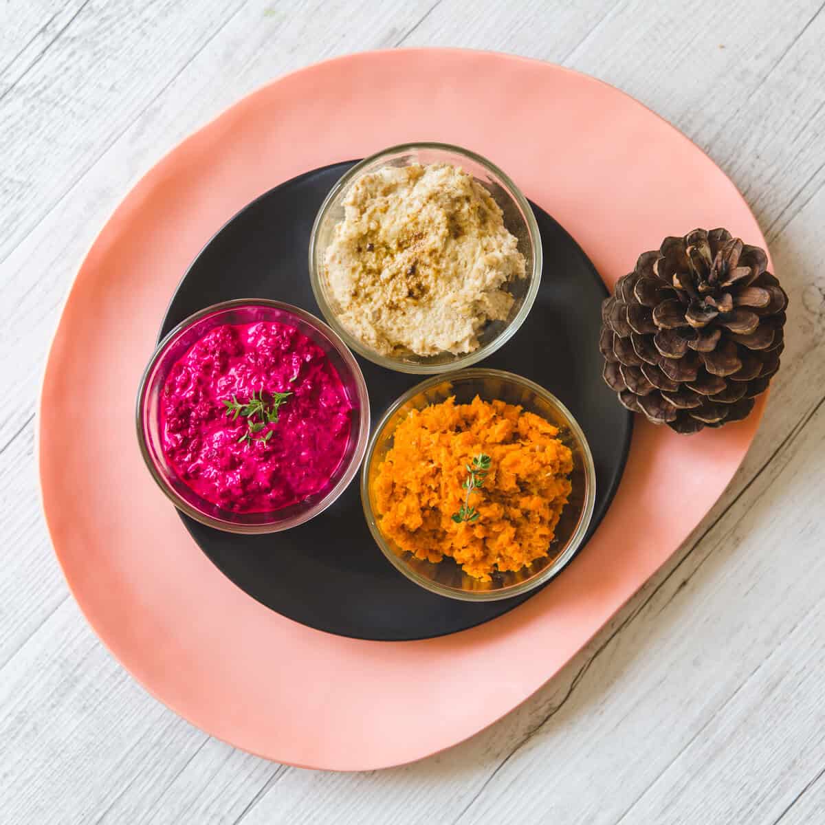Vibrant beetroot dips in glass bowls with pinecone, on pink and black plate.