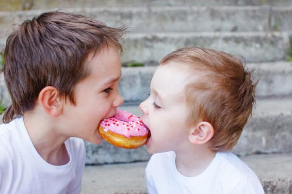 Young boy and toddler sharing a pink frosted donut with sprinkles outdoors.