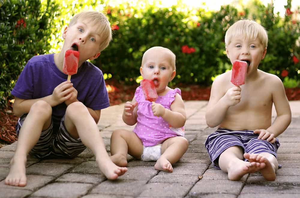 Children sitting on a brick patio eating colourful ice pops on a sunny day.