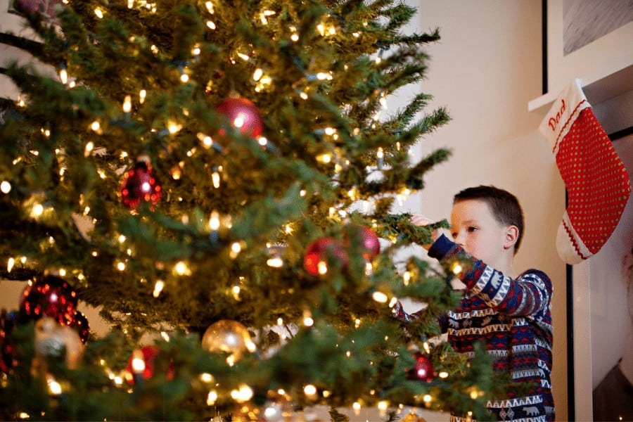 Christmas tree decorated with ornaments and lights, with a young boy reaching for a decoration.