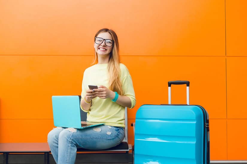 Young woman using smartphone at airport with luggage and laptop.