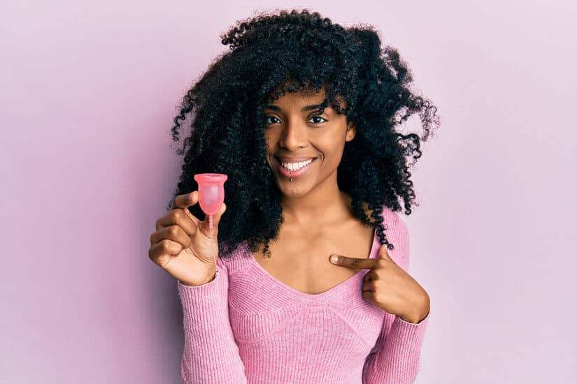 Menstrual cup held by a smiling woman with curly hair, pointing to it, on a pink background.