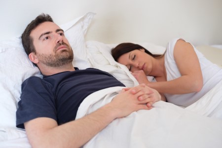 Couple lying in bed, man looking away, woman sleeping peacefully, relationship issues.