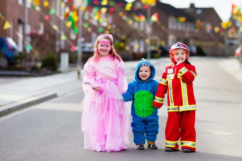 Kids dressed as a princess, firefighter, and superhero enjoying a festive street event.