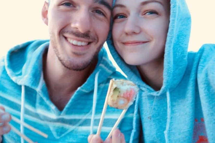 Happy couple sharing sushi outdoors in casual hoodies, smiling at the camera.