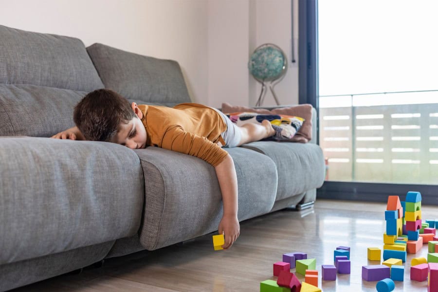 Child lying on a grey sofa reaching for colorful building blocks on the floor.