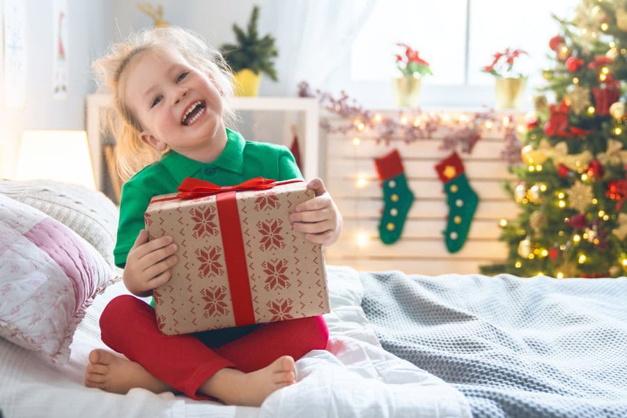 Happy girl holding a wrapped Christmas gift in a festive living room setting.
