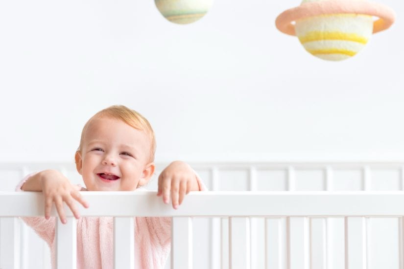Adorable baby smiling in a white crib with colorful hanging decorations above.