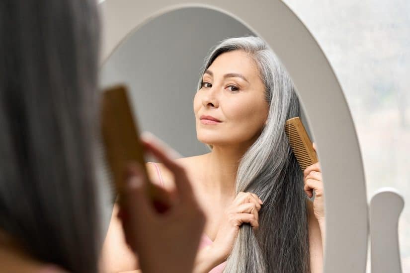 Woman with long, grey hair brushing her hair in front of a mirror.