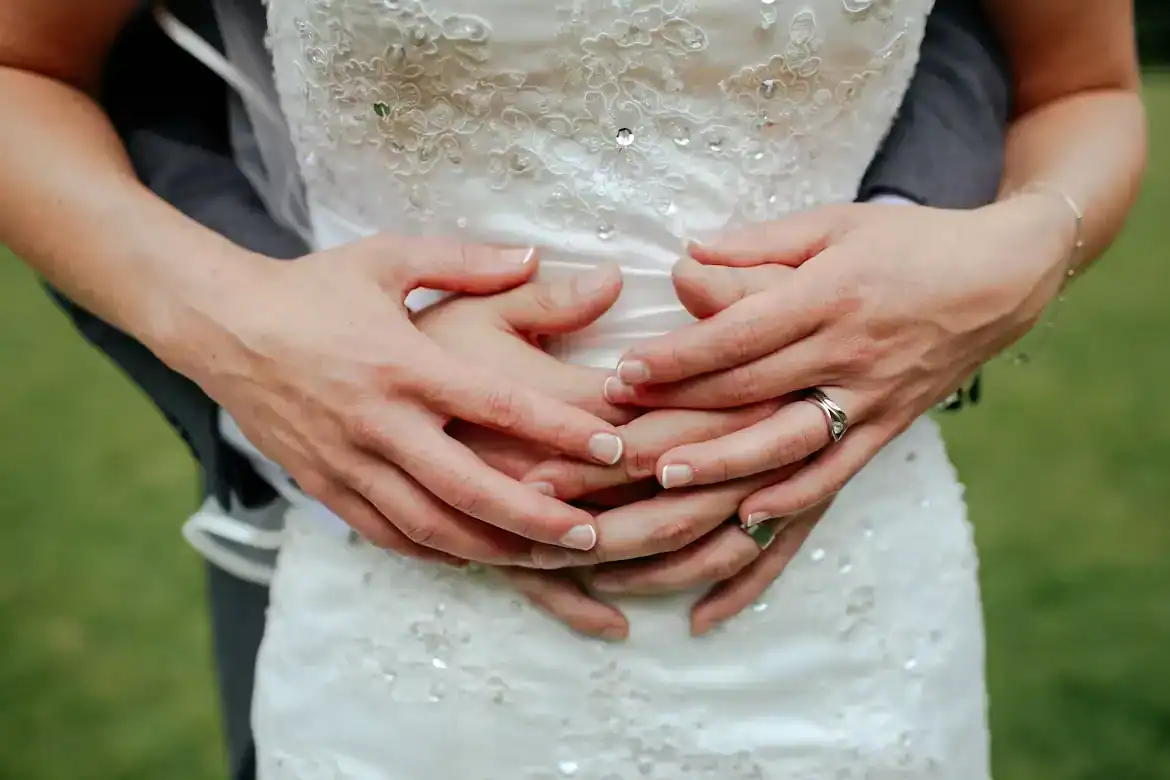 Pregnant bride and partner's hands on her belly, symbolizing love and new beginnings.