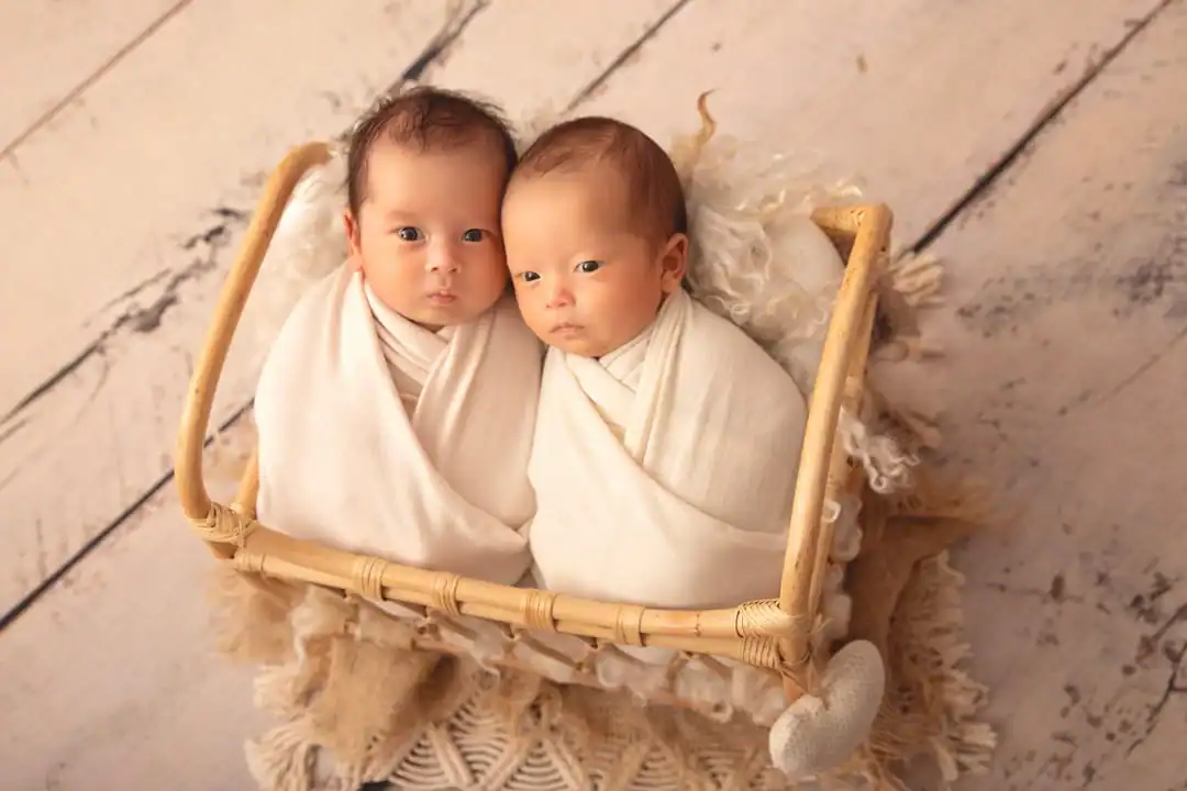 Newborn twins swaddled in cream wraps, lying side by side in a cane bed